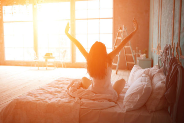 Bettwäsche-Sommer: Frau sitzt auf dem Bett und streckt sich vor einem Sonnenerhelltem Fenster