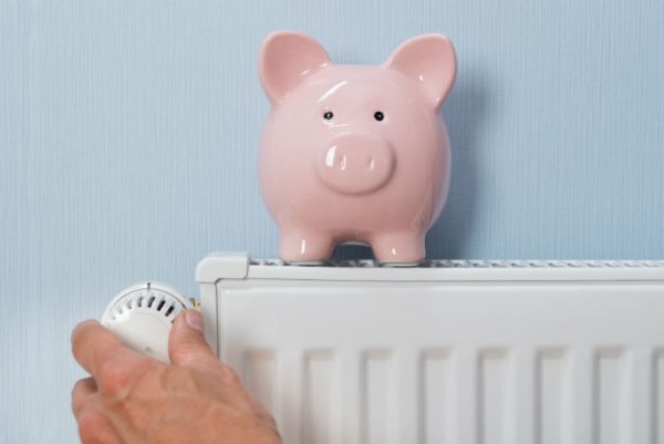 Mann hält Thermostat mit Sparschwein auf Heizkörper